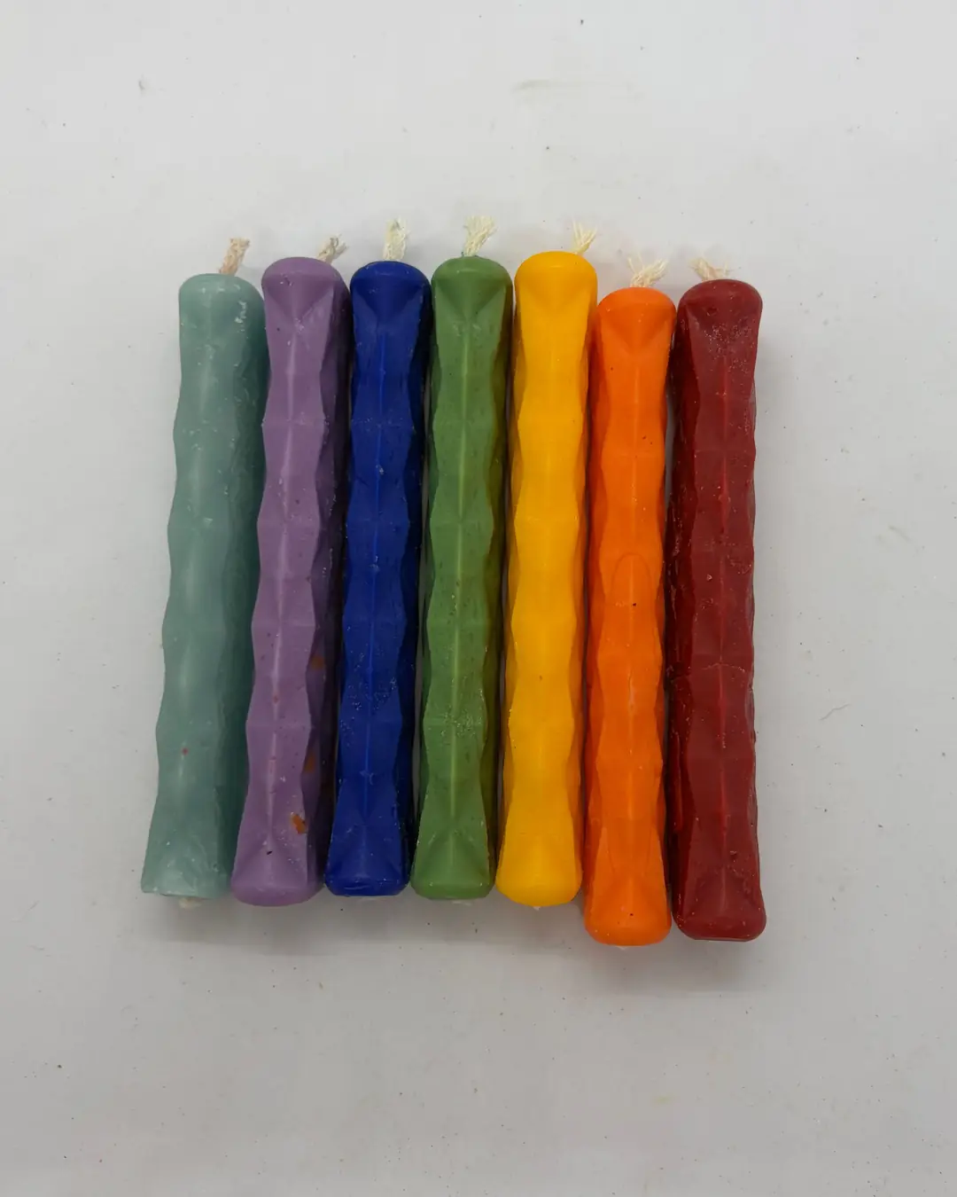 Siete candelas delgadas de cera labrada, una de cada color, alineadas en fila sobre fondo blanco.