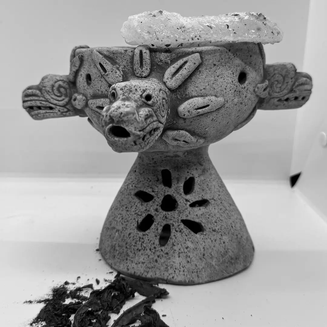 Copalera de barro claro con un trozo de copal encima y astillas de madera al pie.