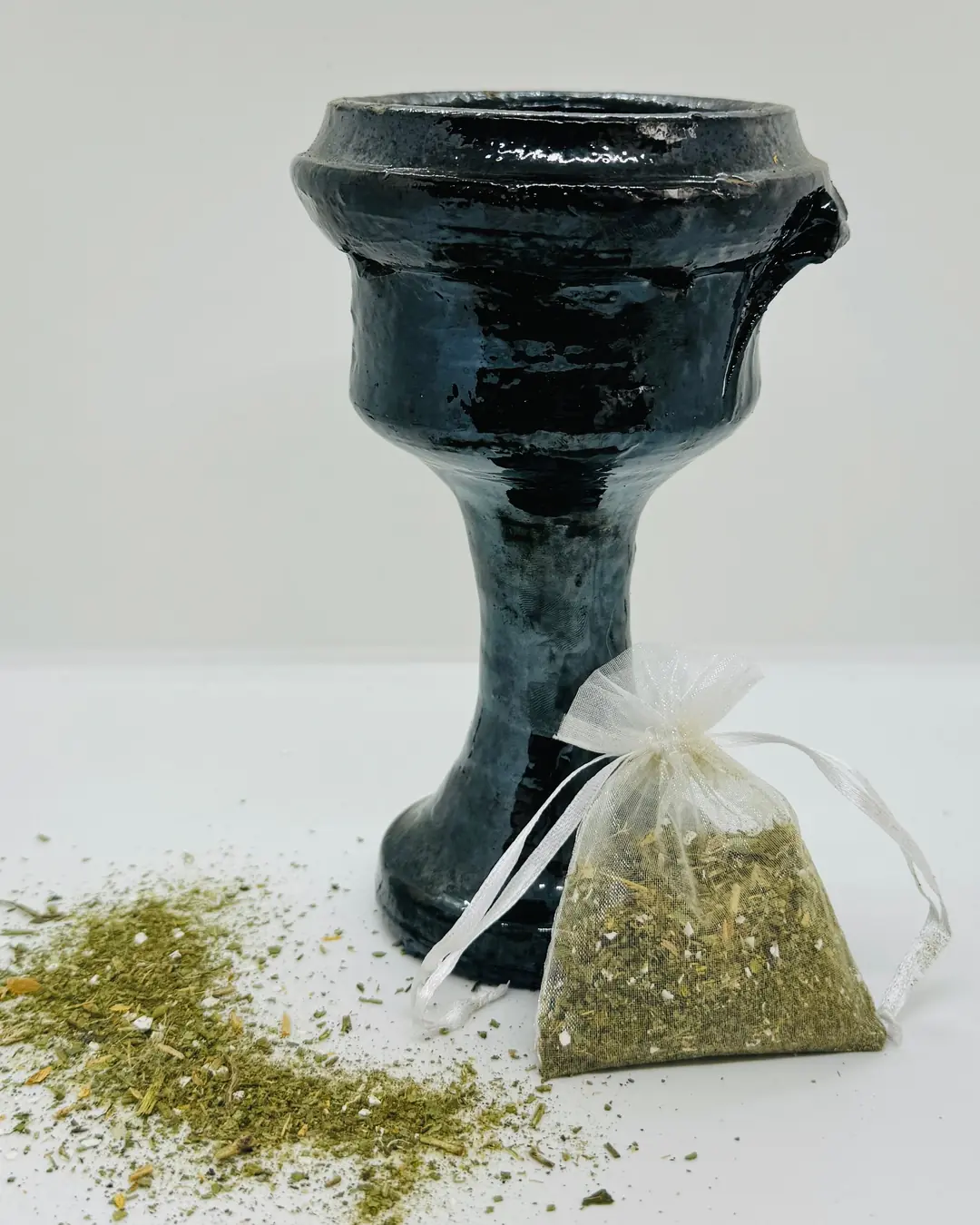Copalera de barro negro vidriado en forma de cáliz, junto a un costal de organza con hierba molida y más hierba esparcida al pie.