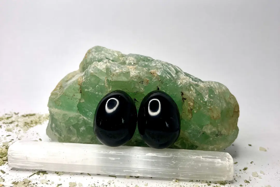 Dos huevos de obsidiana negra pulida frente a un trozo de fluorita verde y una barra de selenita.