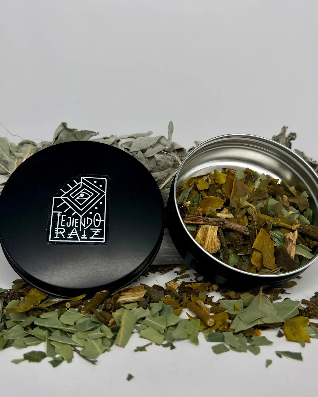Lata abierta con hojas secas de muérdago y astillas, junto a su tapa negra; detrás, salvia seca y hojas esparcidas al frente.