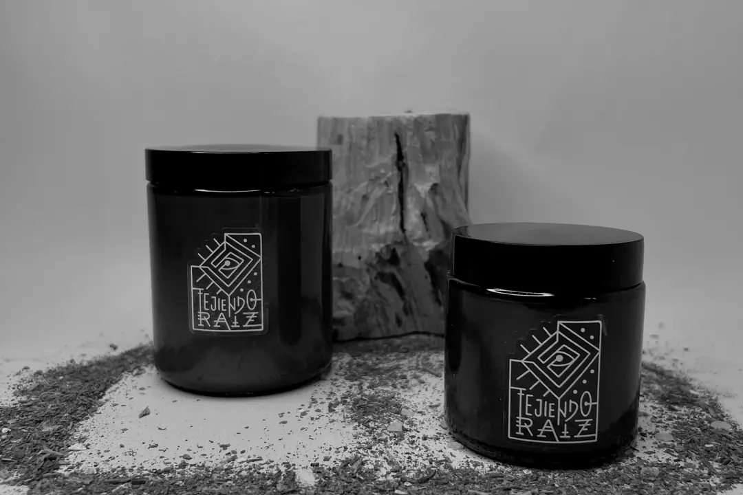 Dos velas en frasco de vidrio ámbar con tapa negra, frente a un tronco de palo santo, sobre hierba seca.
