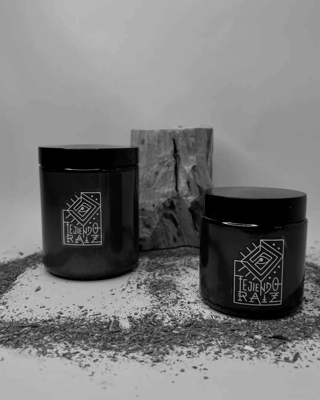 Dos velas en frasco de vidrio ámbar con tapa negra, frente a un tronco de palo santo, sobre hierba seca.
