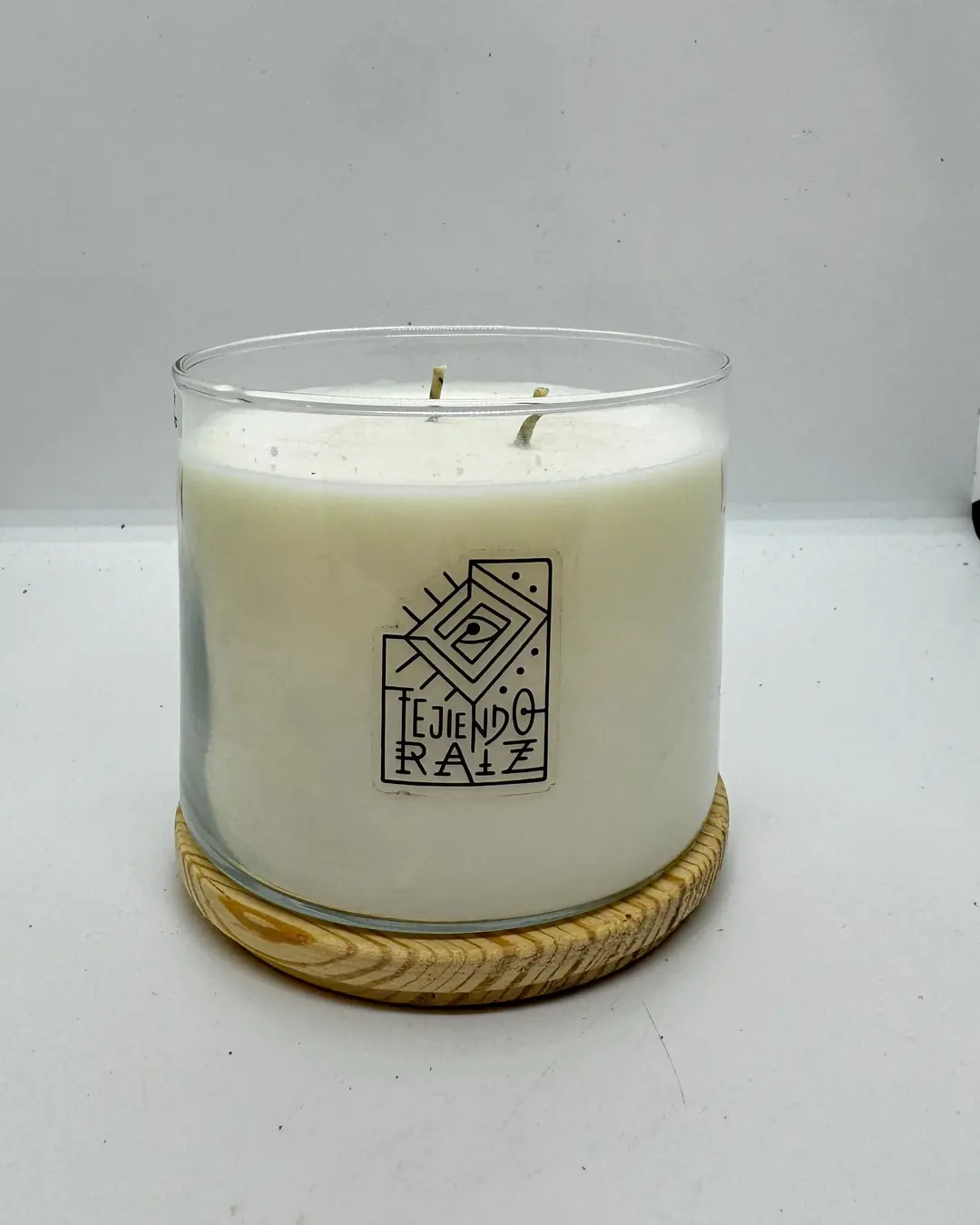 Vela grande de dos pabilos en frasco de vidrio transparente sobre una base redonda de madera, con el logo de Tejiendo Raíz.